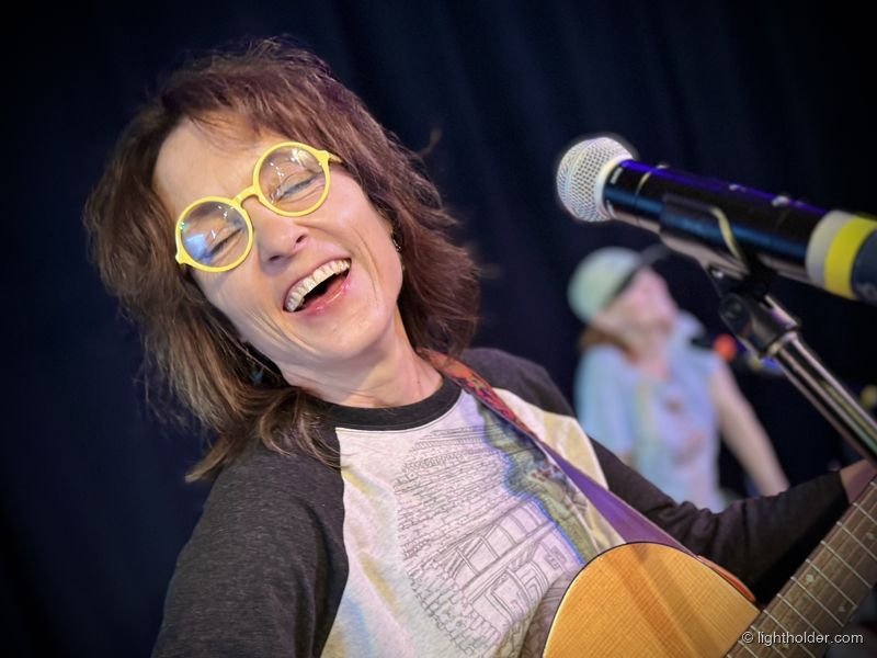 A smiling Deborah Crooks wears yellow-framed bird glasses while playing guitar